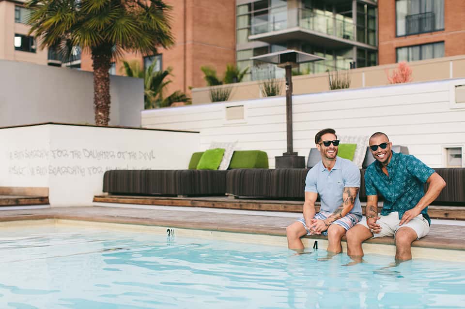 Two men relaxing poolside at an urban hotel courtyard with palm trees and white lounge seating