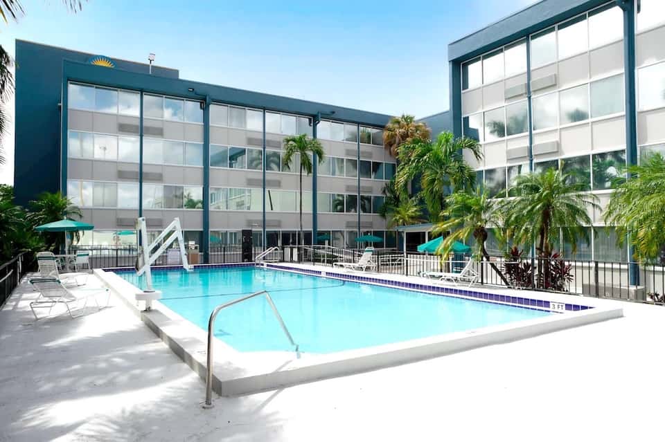 Hotel pool surrounded by white lounge chairs, palm trees, and multi-story residential building