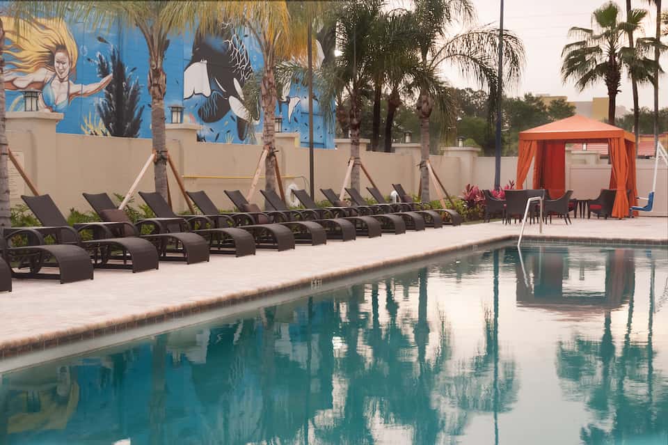 Resort pool deck with black loungers, blue mural wall, orange cabana, and palm trees