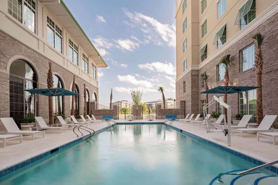 Modern hotel courtyard pool with arched windows, white loungers, blue umbrellas, and palm trees