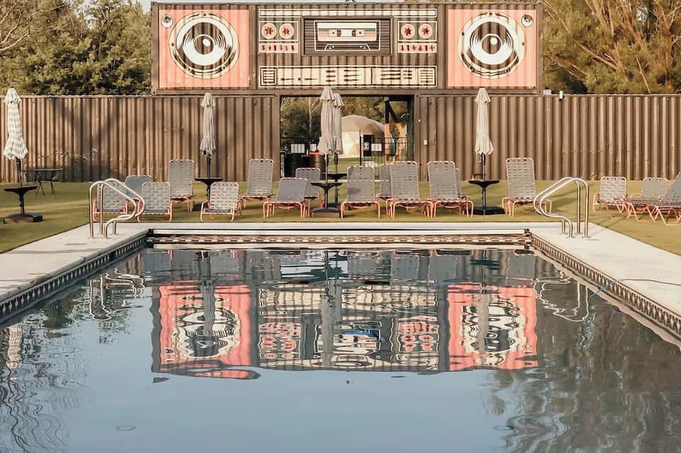 Retro-style pool with pink and white boombox art installation, lawn chairs, and corrugated metal backdrop