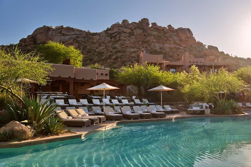 Resort pool with turquoise water, loungers, umbrellas, and rocky mountain backdrop at sunset