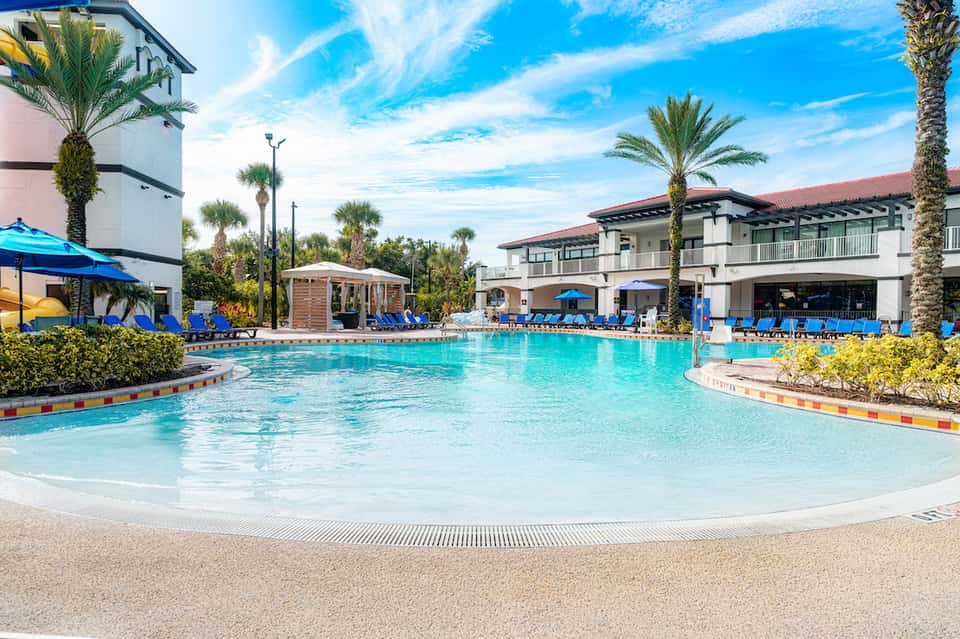 Resort-style pool with turquoise water, palm trees, blue lounge chairs, and white pergolas under clear blue sky