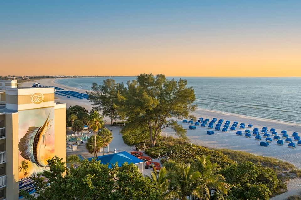 Beachfront resort at sunset with blue umbrellas on sand, ocean view, and illuminated building with tarpon mural