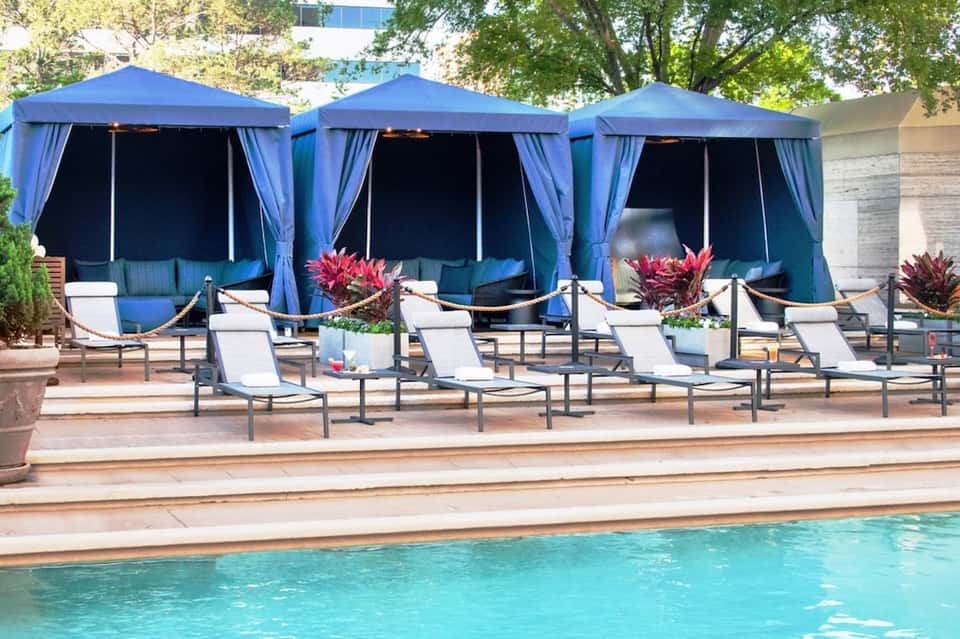 Poolside lounge area with blue cabanas, white loungers, and tropical plants by the water