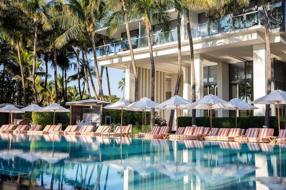 Waterfront resort pool with striped lounge chairs, white umbrellas, palm trees, and modern architecture