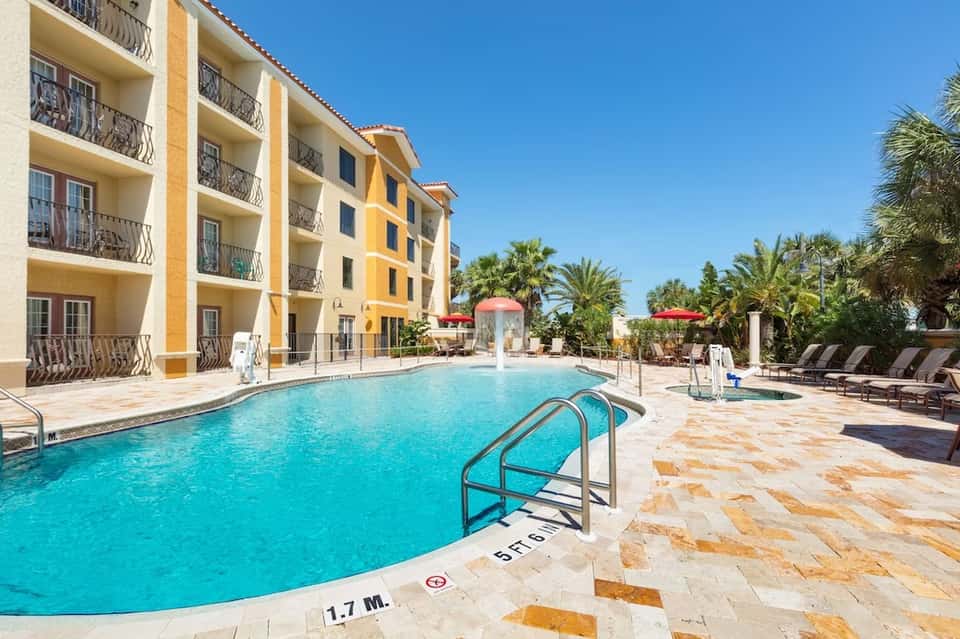 Resort pool with yellow and beige building, palm trees, lounge chairs, and clear blue water
