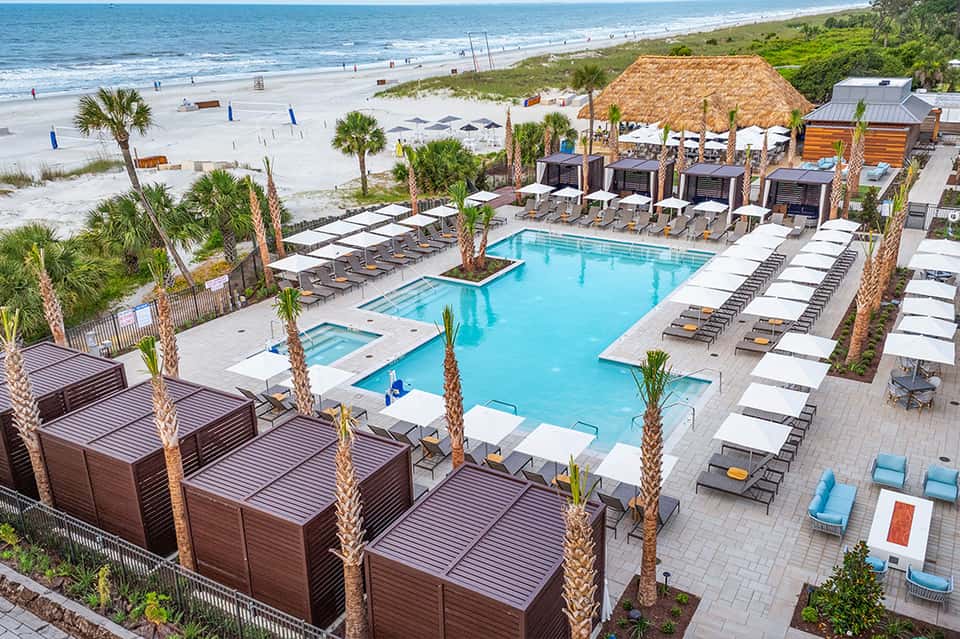 Beach resort pool area with palm trees, cabanas, thatched-roof bar, and ocean view in background