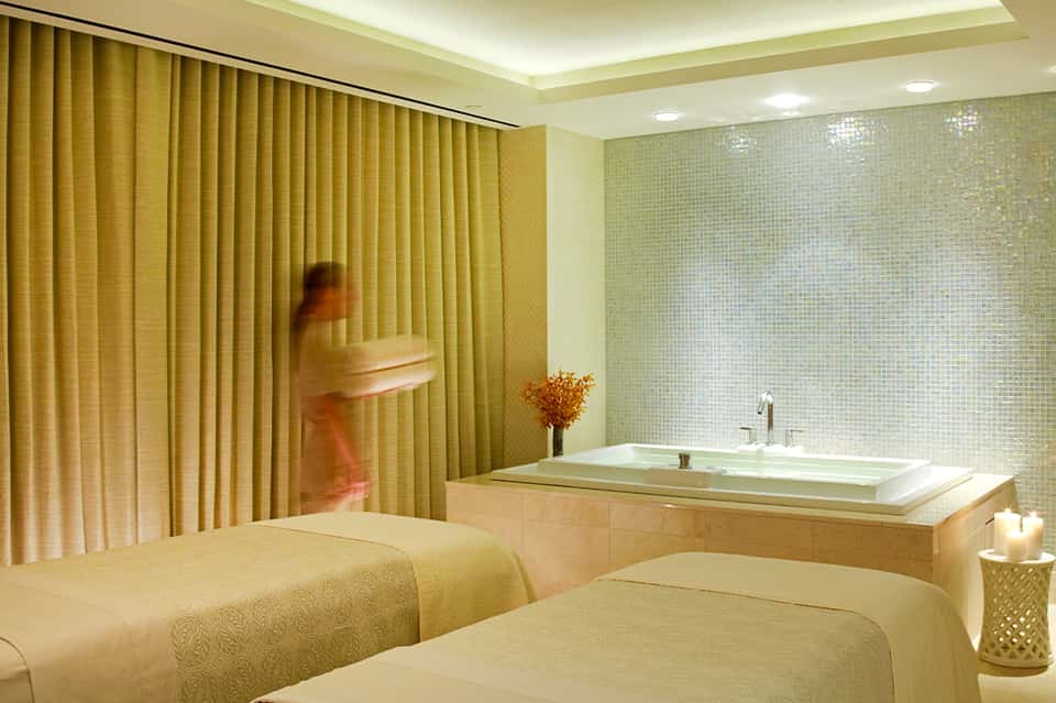 Spa treatment room with massage beds, golden curtains, green tile walls, and soaking tub