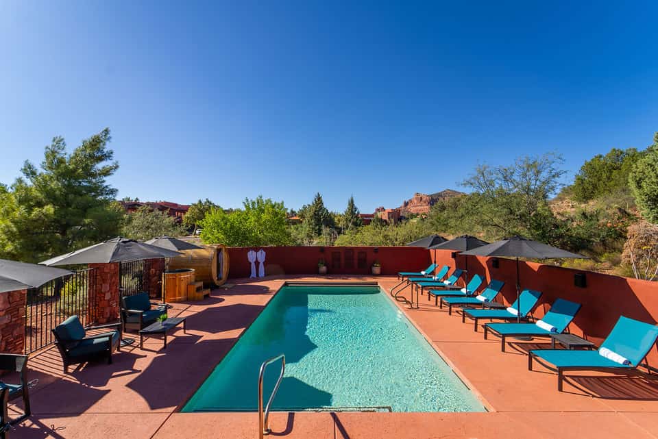 Rectangular turquoise pool on terrace with blue lounge chairs, black umbrellas, and red rock formations in background