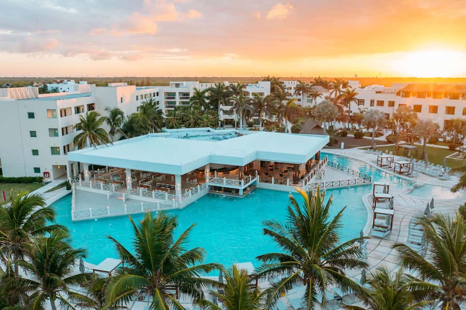 Aerial view of turquoise pool with bar pavilion at sunset, palm trees surrounding