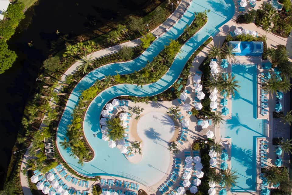 Aerial view of resort with curved lazy river, multiple pools, and white umbrellas among palm trees