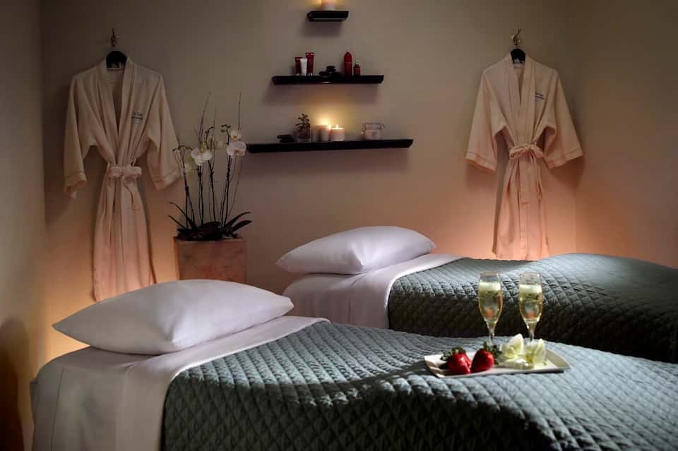 Romantic spa bedroom with two beds, champagne glasses, fresh berries, plush robes, and ambient lighting from wall shelves