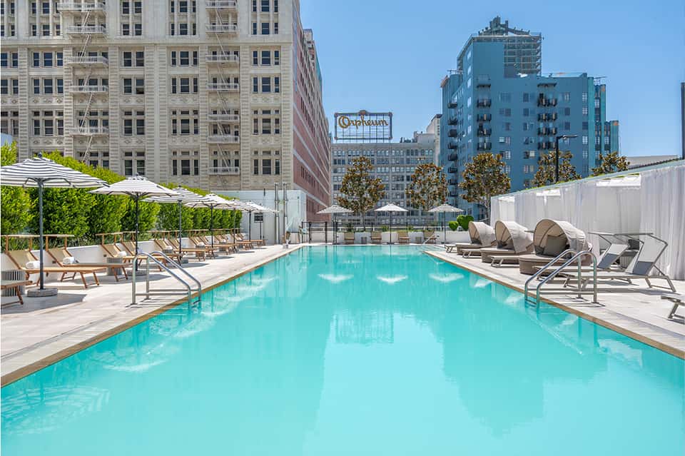 Rooftop pool with turquoise water, lounge chairs, white umbrellas, and urban cityscape backdrop