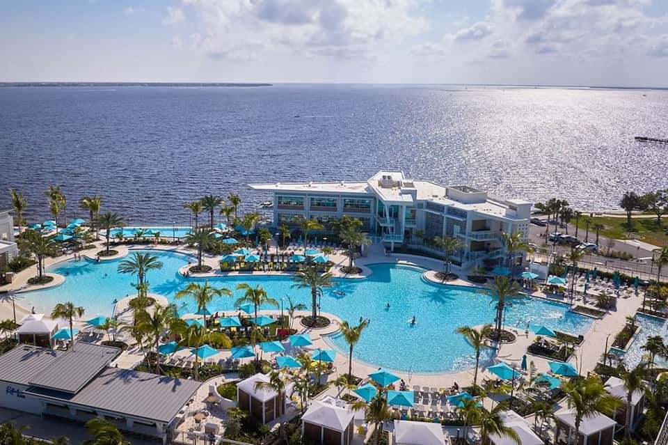 Aerial view of large resort complex with multiple pools, palm trees, and waterfront location