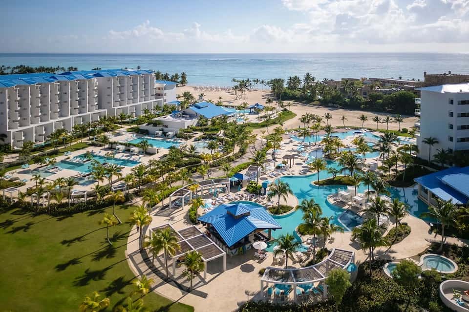 Beachfront resort aerial view with multiple pools, palm trees, white buildings, and ocean