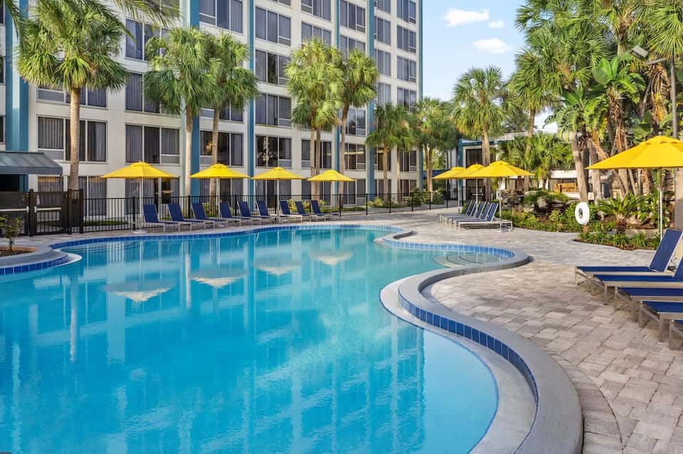 Resort pool area with bright blue water, yellow umbrellas, palm trees, and multi-story hotel building in background