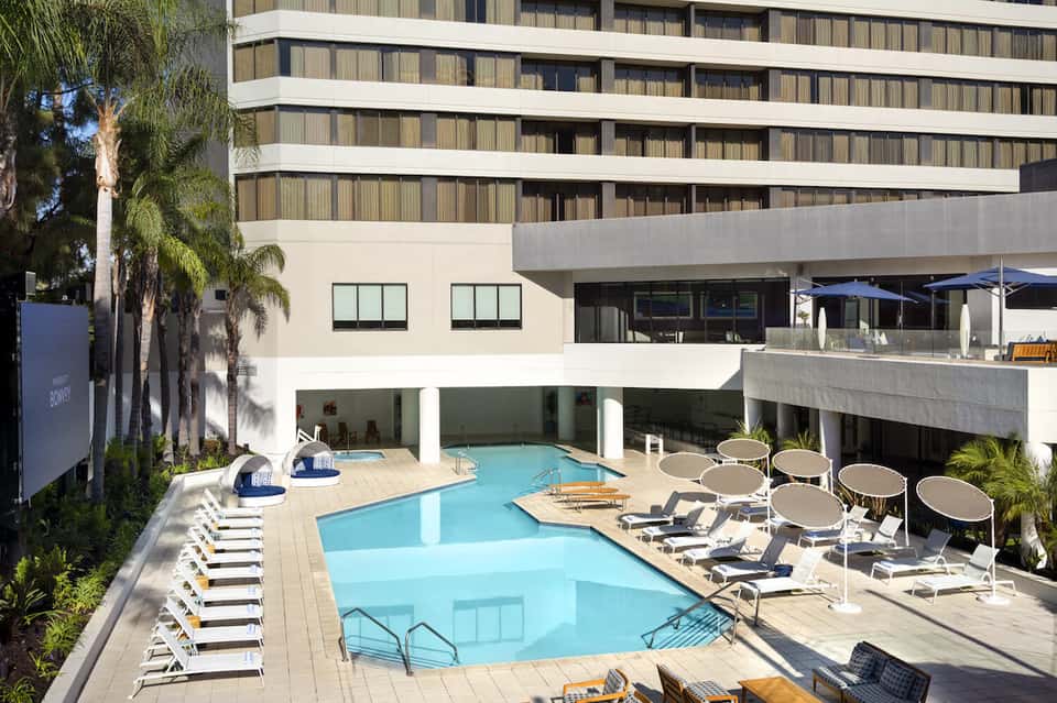 Contemporary hotel pool deck with loungers, umbrellas, and multi-story building surrounded by palm trees