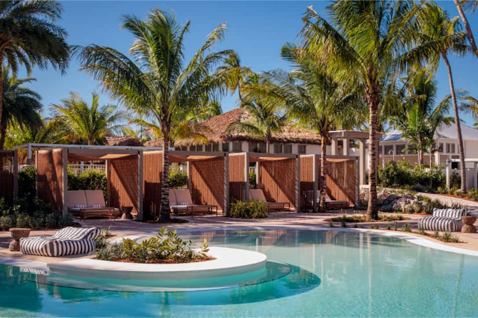 Resort pool with turquoise water, wooden cabanas, palm trees, and thatched-roof structures