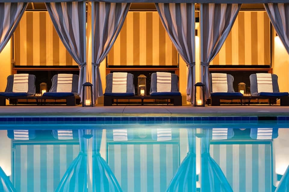 Elegant poolside cabanas at dusk with striped loungers, draped curtains, and golden lighting