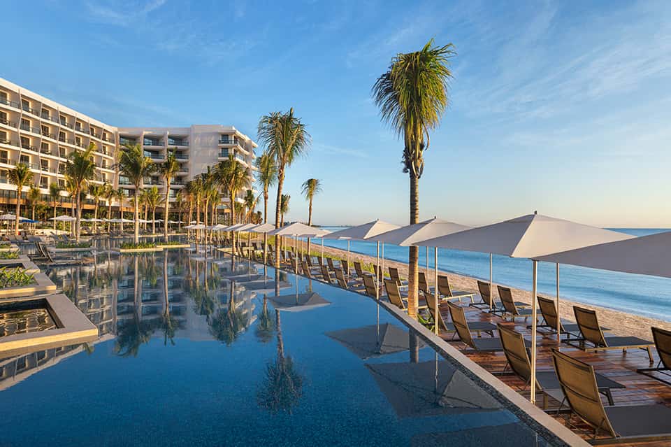 Beachfront resort pool with reflective water surface, palm trees, loungers, shade structures, and ocean view