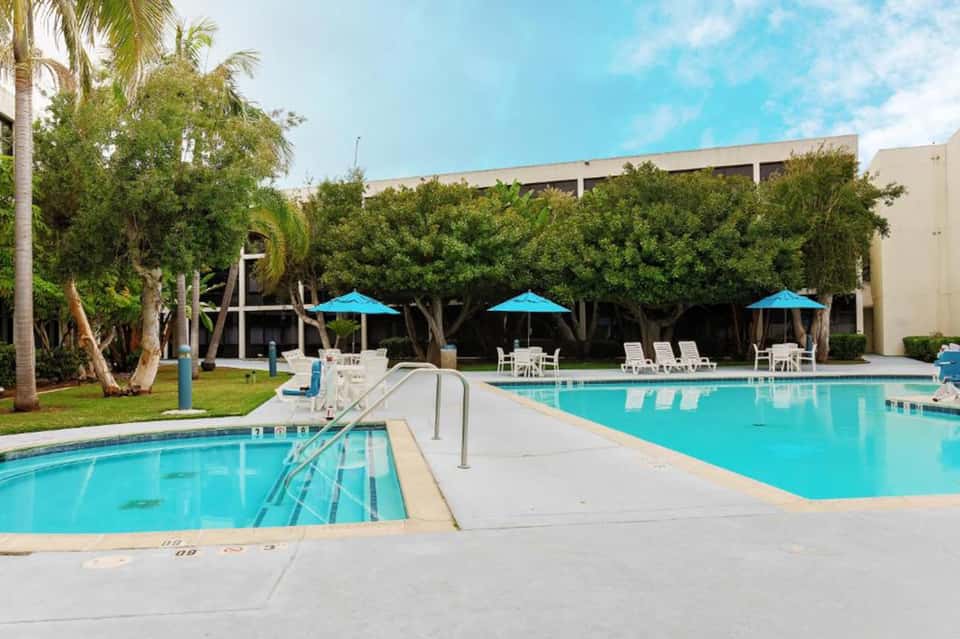 Two connected outdoor pools with lounge chairs, blue umbrellas, mature trees, and building backdrop