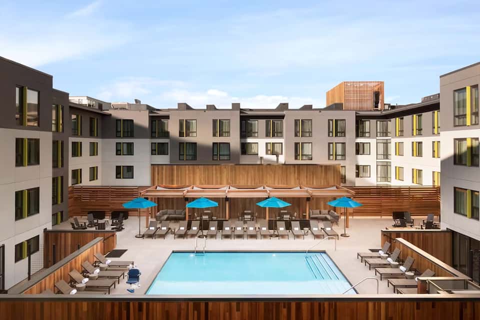 Urban rooftop pool surrounded by modern apartment buildings with blue umbrellas and loungers