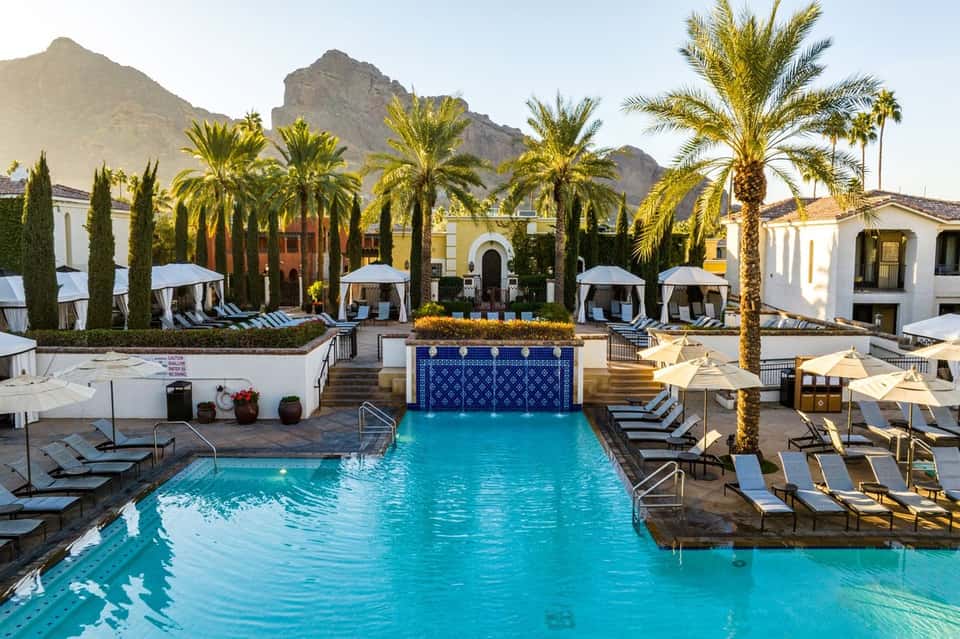 Resort pool courtyard with turquoise water, palm trees, white umbrellas, and mountain backdrop