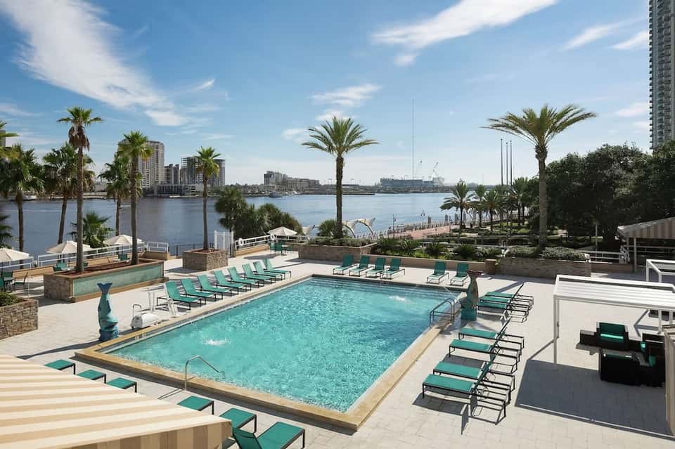 Waterfront pool with turquoise water, lounge chairs, palm trees, and city skyline views