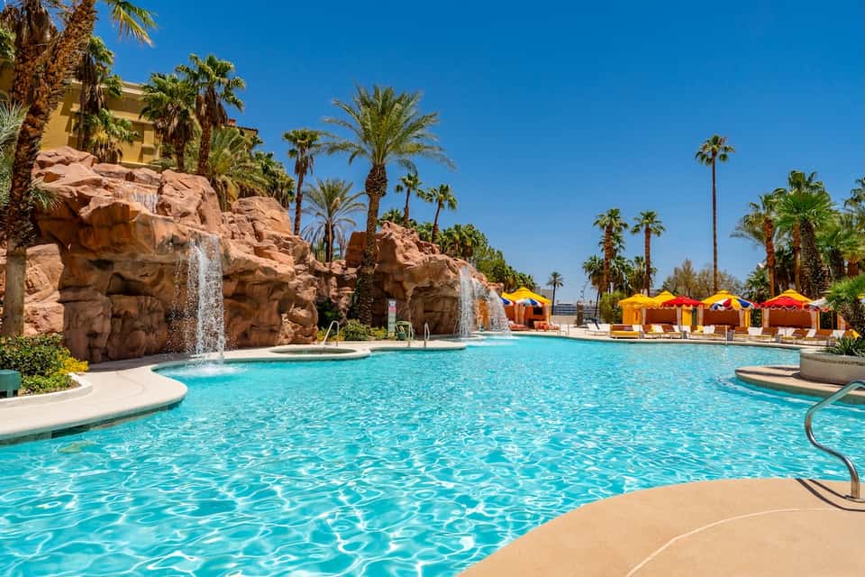Resort pool with turquoise water, rocky waterfall features, palm trees, and yellow cabanas
