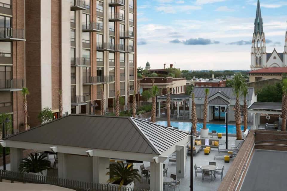 Elevated view of resort pool deck with white pergola, palm trees, surrounding hotel buildings, and distant church spire