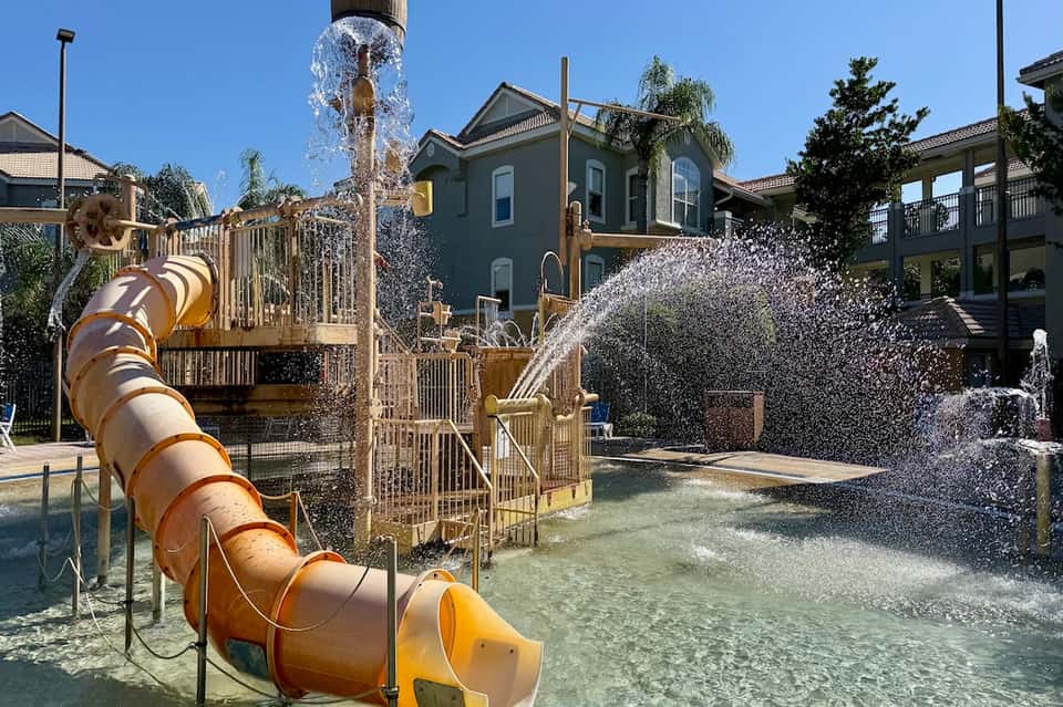 Water park splash pad area with bronze slide, water fountains, and children's play structures in shallow water
