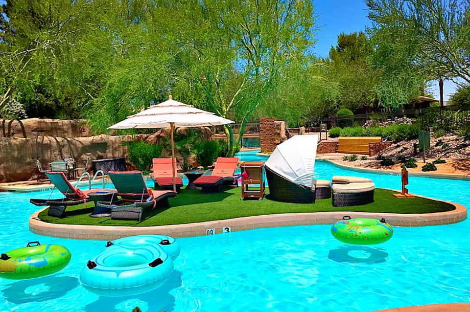 Resort pool with turquoise water, lounge chairs, umbrellas, and lush green landscaping surrounded by willow trees