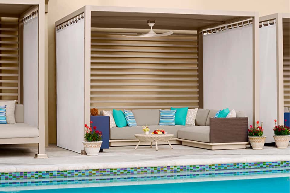 Resort pool area with striped pergola canopy, cushioned lounge seating, potted flowers, and motorized shade screens.