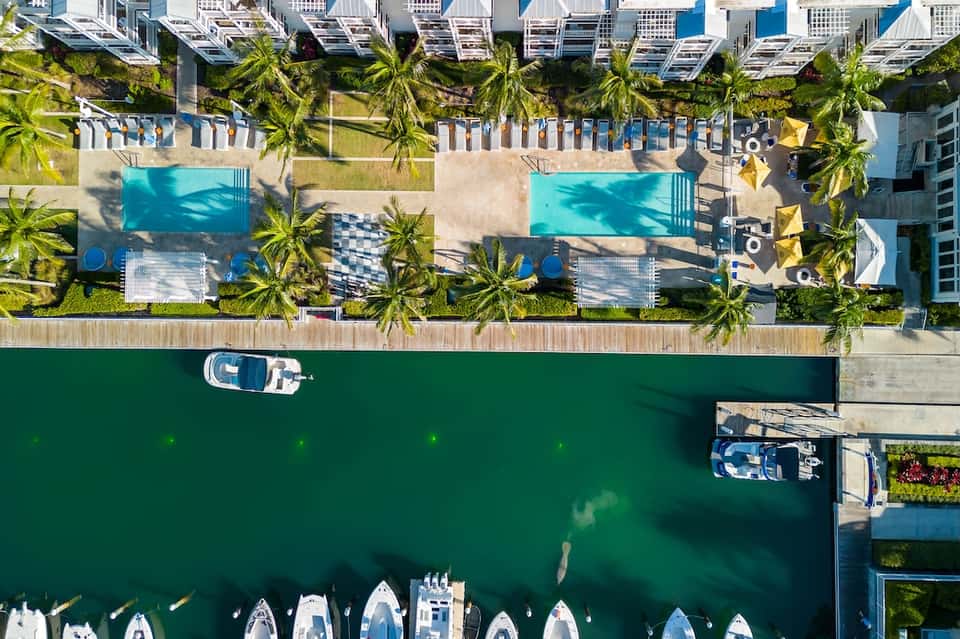 Overhead view of waterfront resort with pools, loungers, palm trees, boats, and green waterway