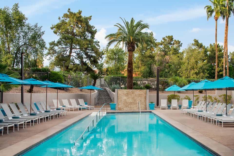Resort swimming pool with turquoise water, white lounge chairs, blue umbrellas, and palm tree landscape