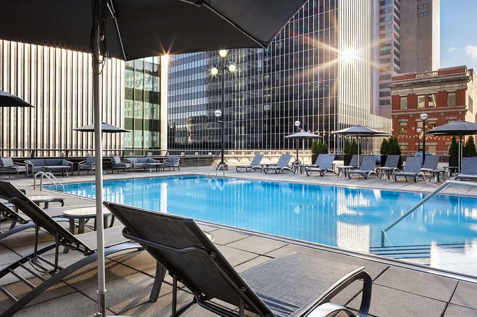 Rooftop pool deck at urban hotel with blue water, black lounge chairs, umbrellas, and city skyline views