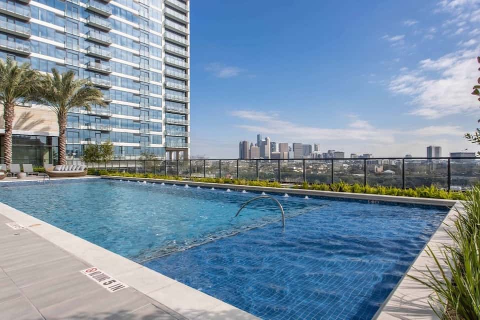 High-rise rooftop pool with city skyline view, palm trees, and lounge chairs along the deck edge