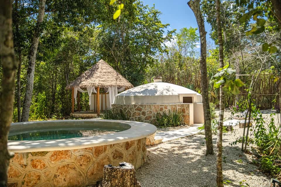 Tropical wellness area with natural hot spring pool, thatched hut, and stone structures surrounded by forest