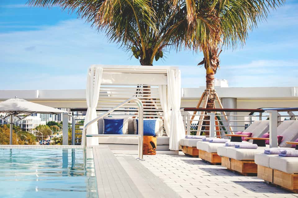 Luxury poolside cabana with white curtains, palm tree, and loungers overlooking swimming pool