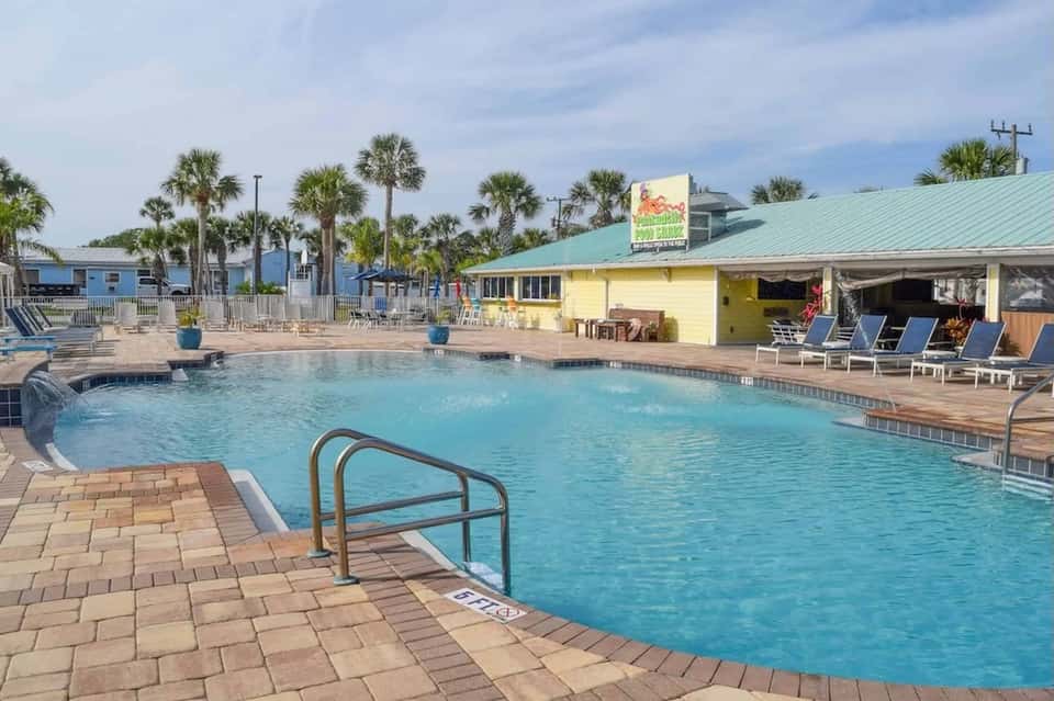 Resort pool with yellow cabana building, lounge chairs, and palm trees