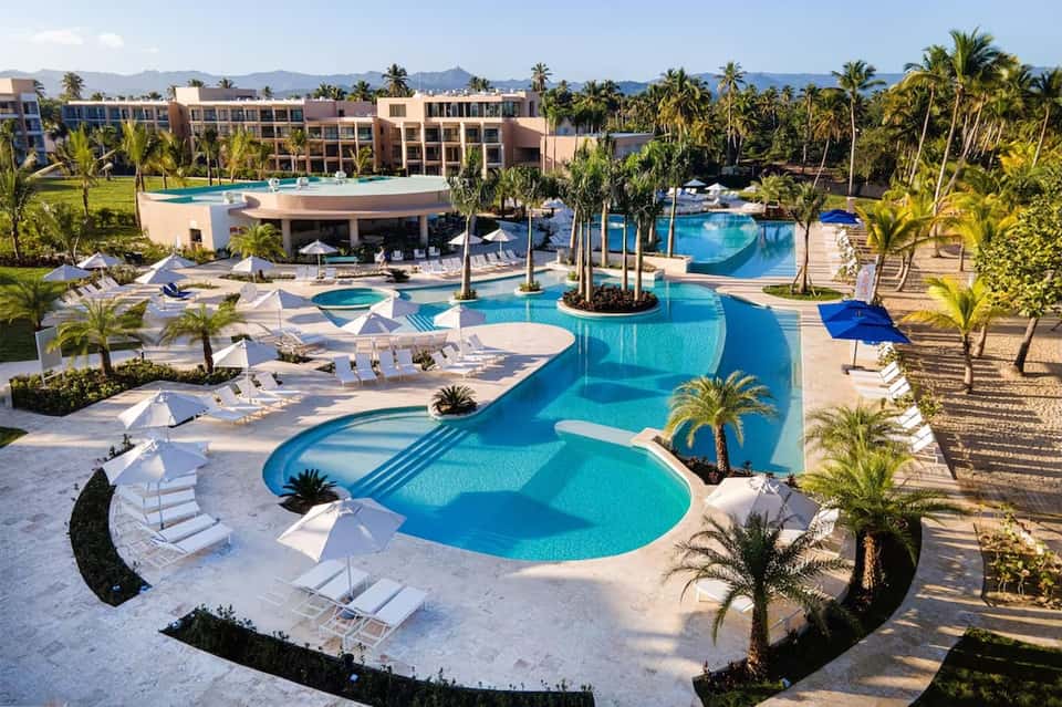 Aerial view of sprawling resort with multiple pools, palm trees, and mountains