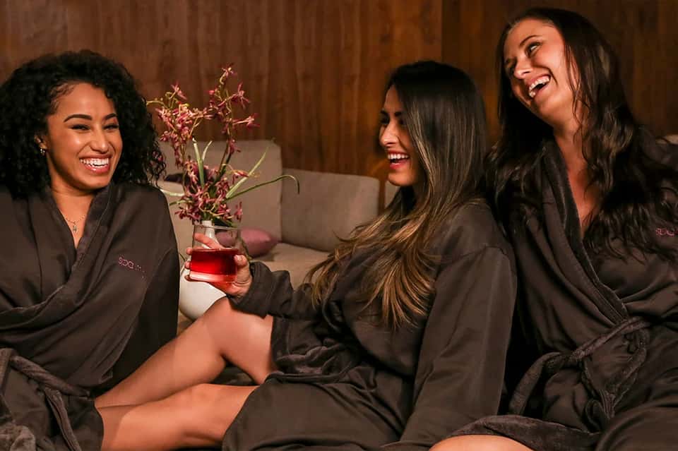 Three women in spa robes laughing together in a lounge area with flowers