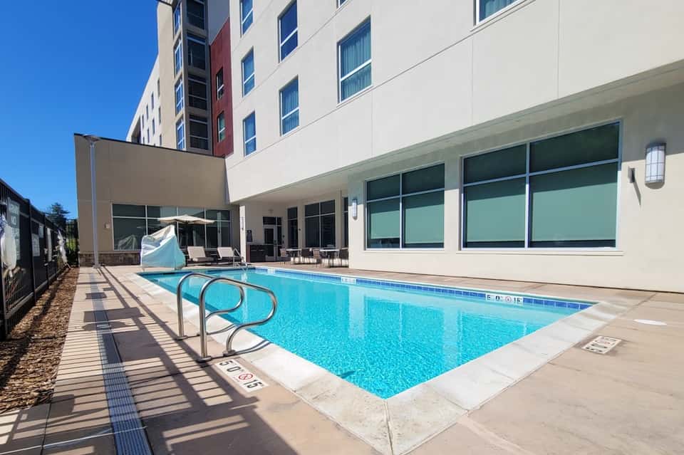 Modern hotel pool with blue water, deck chairs, shade sail, and contemporary building facade