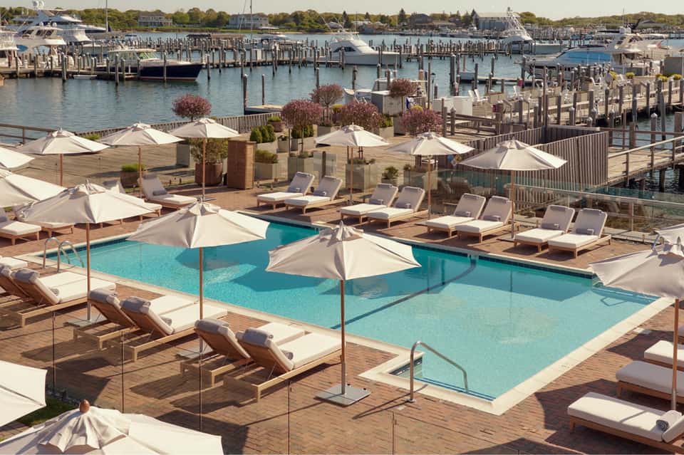 Waterfront pool deck with turquoise water, lounge chairs, white umbrellas, and marina views in background
