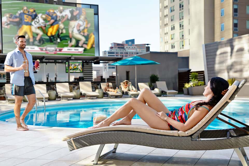 Rooftop pool area with woman lounging, man standing nearby, sports broadcast on large screen, city skyline background