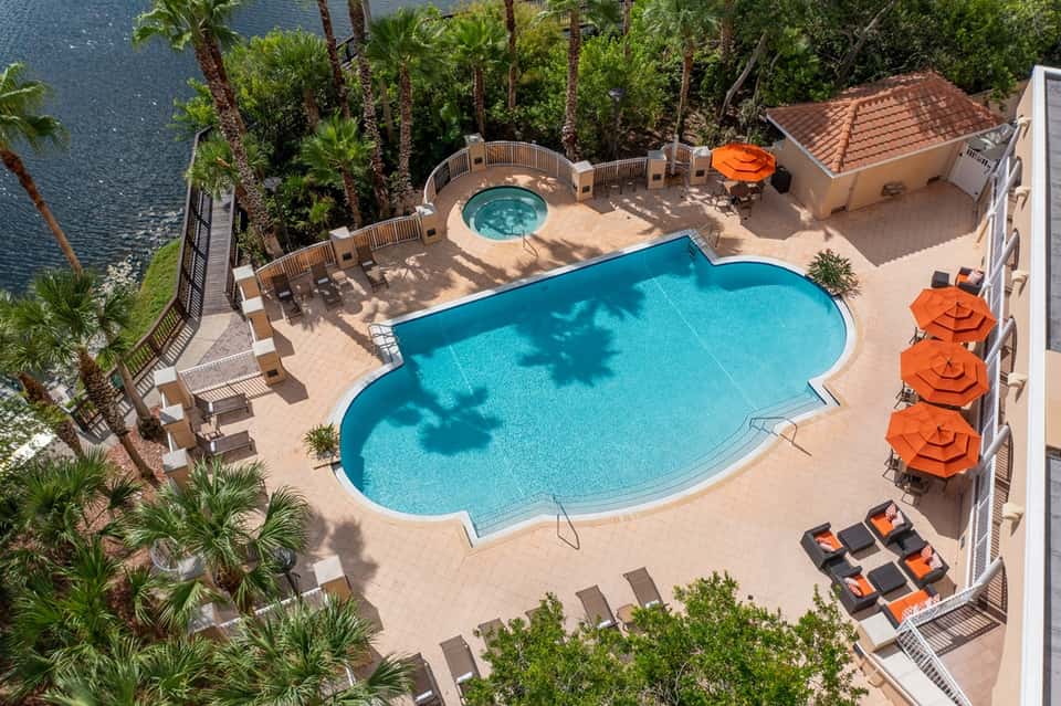 Aerial view of resort pool with spa tub, loungers, orange umbrellas, and waterfront palm trees