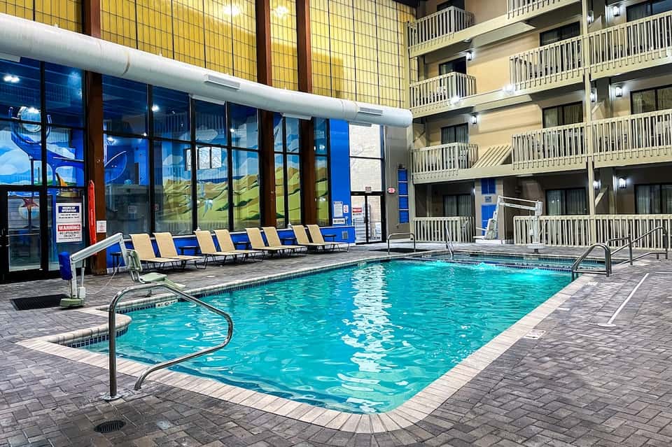 Indoor pool with blue and yellow loungers, large windows, and multi-story hotel building overlooking
