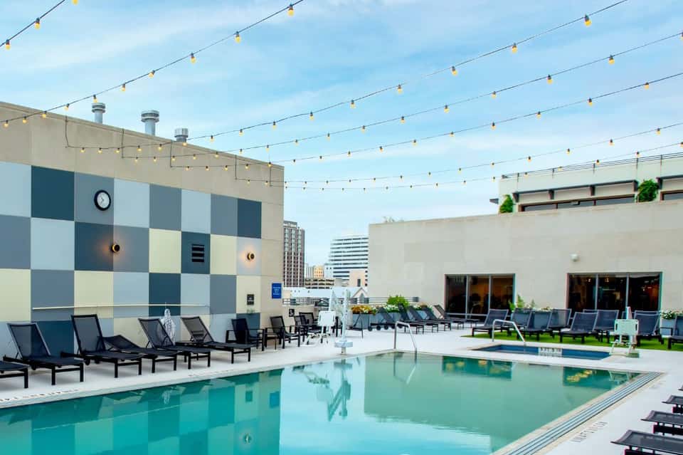 Modern rooftop pool with string lights, lounge chairs, and city skyline views during daytime