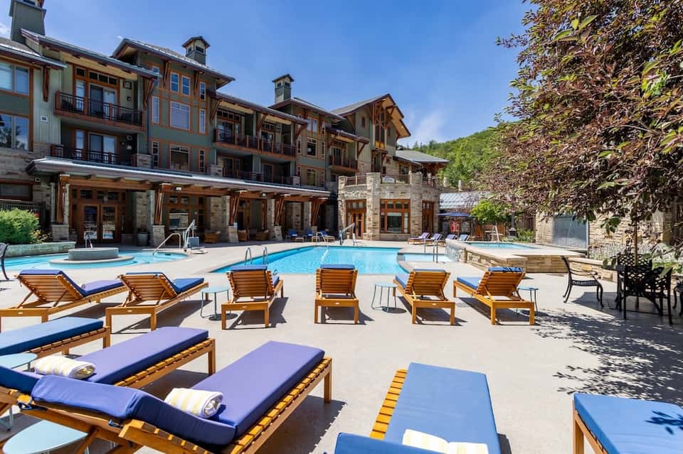 Resort pool with wooden lounge chairs, blue cushions, and multi-story lodge building